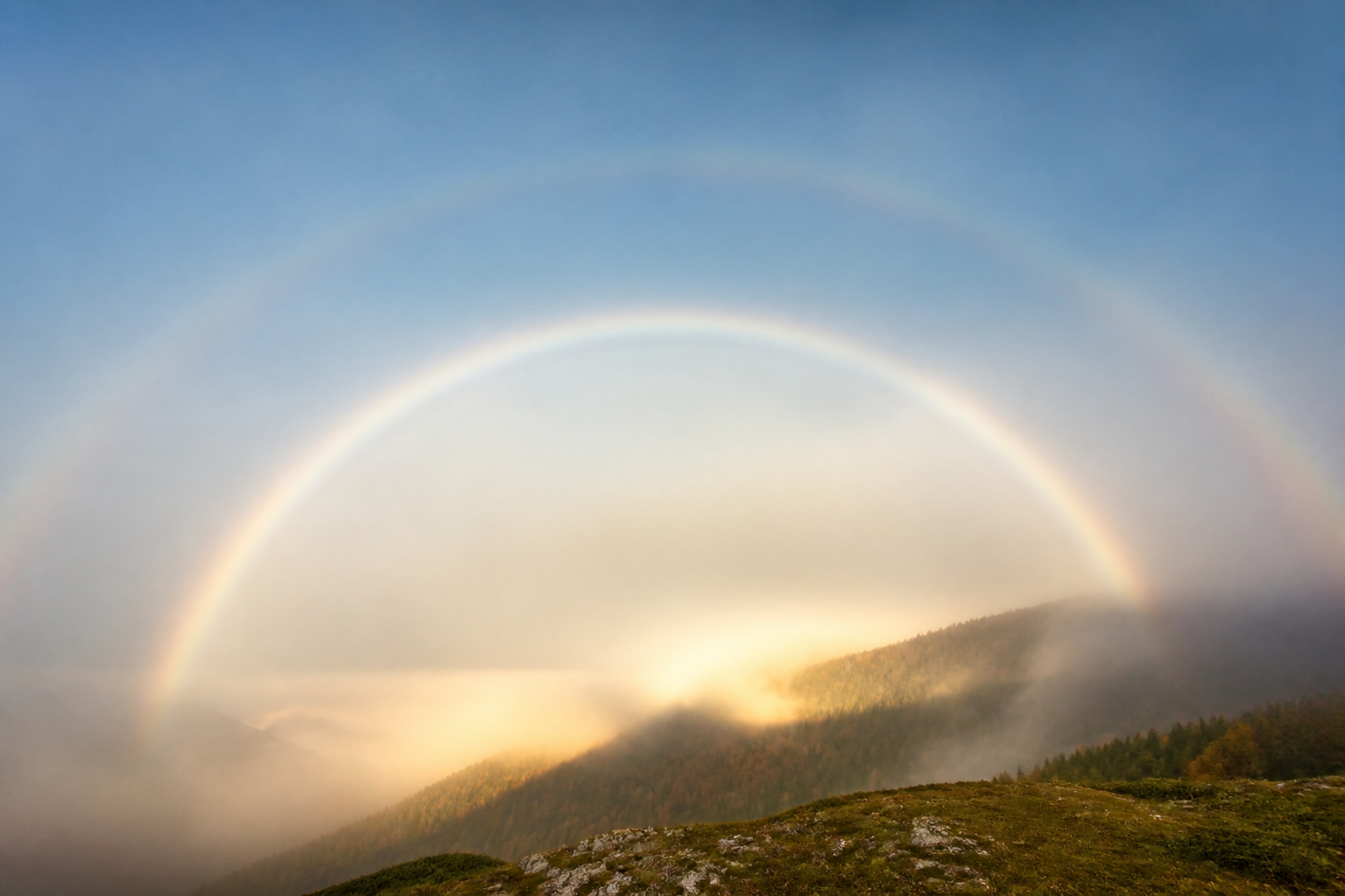 Arc blanc large et pâle se détachant dans une nappe de brouillard éclairée par le soleil bas