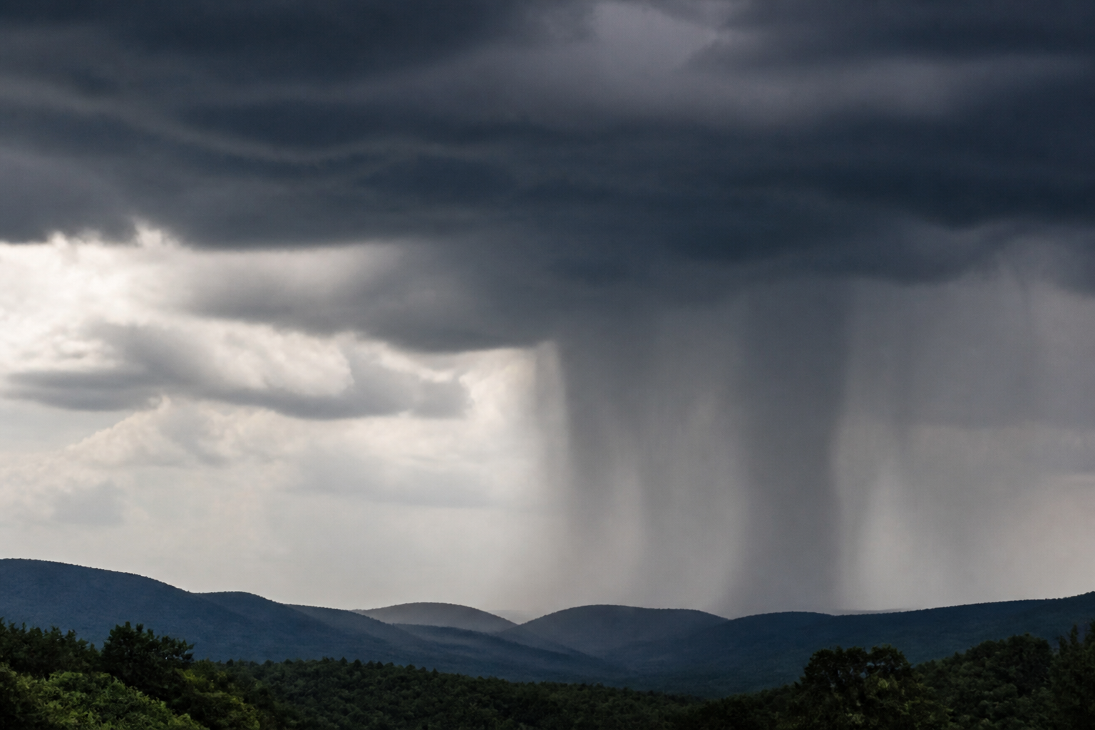 Rideau de pluie sombre tombant verticalement sous la base d'un cumulonimbus, éclairé de côté par le soleil